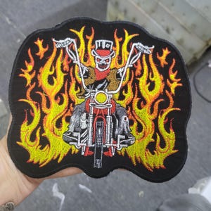 May include: Embroidered patch featuring a skeleton biker riding a red motorcycle, surrounded by flames in shades of yellow and orange. The patch has a black border and is designed with detailed stitching.