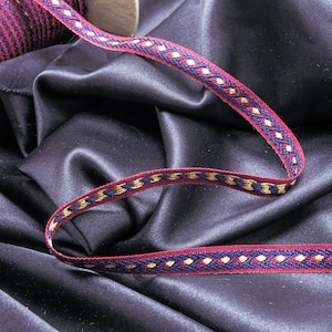 May include: A close-up of a burgundy and gold ribbon with a blue and gold geometric pattern. The ribbon is lying on a dark blue fabric.