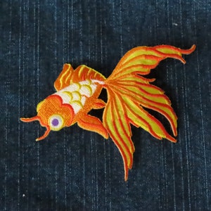 Peut inclure: Un écusson brodé thermocollant représentant un poisson rouge orange vif avec des nageoires jaunes et rouges. Le poisson rouge a un corps blanc avec des écailles orange et jaunes.