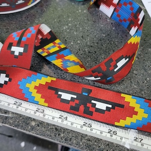 Puede incluir: Cinta roja estampada con un diseño pixelado en negro, blanco, azul y amarillo. Se coloca una regla junto a la cinta. La cinta tiene un patrón geométrico repetitivo.