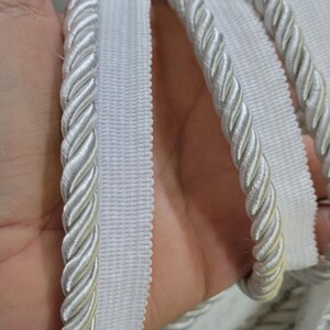 May include: A white decorative trim featuring a twisted rope cord attached to a flat, woven fabric tape edge. The material has a subtle sheen and a uniform, clean appearance, designed for sewing applications and textile finishing projects.