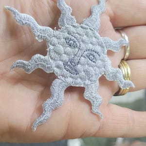 May include: A silver-colored embroidered patch featuring a sun design with a central face. The sun has a circular center with a stitched facial expression and multiple wavy, pointed rays extending outward from the perimeter of the circle.
