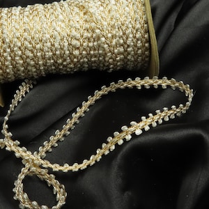 Op de afbeelding: Een rol decoratieve sierband in crème- en goudtinten. De band, met een getextureerde afwerking, wordt getoond op zwarte satijnen stof. Geschikt voor naai- en knutselprojecten, voegt het een decoratief tintje toe.