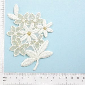 May include: White lace flower applique with gold accents. The applique is approximately 6 cm wide and 5 cm tall.