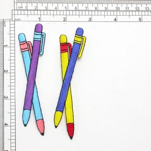 May include: Three embroidered iron-on patches of pens in blue, purple, and yellow with red and pink accents.
