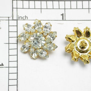 May include: Two gold-toned flower-shaped buttons with clear rhinestone accents. The button on the left has a clear rhinestone center, while the button on the right has a gold center.