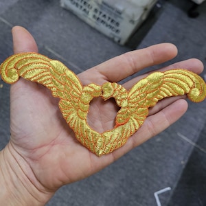 May include: Embroidered gold-colored wings with orange accents, forming a heart shape in the center. The wings feature detailed stitching and a textured appearance, suitable for apparel or craft projects.