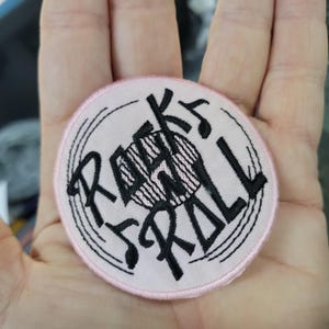 Könnte beinhalten: Ein runder, hellrosa Aufnäher mit dem schwarzen Schriftzug "ROCK 'N' ROLL". Der Aufnäher hat einen schwarzen Rand und Noten. Die Mitte des Aufnähers hat ein stilisiertes Design.