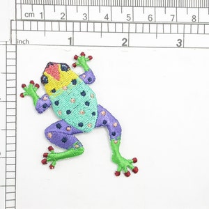 May include: A colourful embroidered frog patch with green, purple, yellow and red accents. The frog has a spotted pattern and is approximately 5 cm in length.