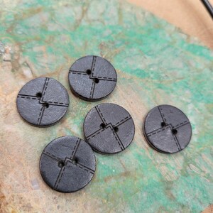 May include: A collection of five dark gray, round buttons with a stitched design. Each button has two holes and a cross-stitched pattern, creating a textured appearance. The buttons are scattered on a green and brown surface.