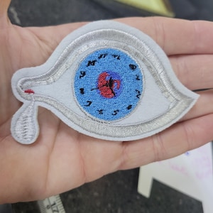 Op de afbeelding: Geborduurde patch in de vorm van een oog met een wijzerplaat. Het oog is wit met een zilveren rand en een blauwe wijzerplaat met zwarte cijfers. Een rode traan is geborduurd aan de onderkant van het oog.