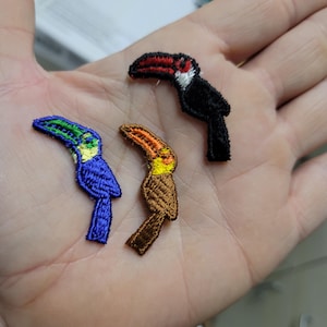 May include: Embroidered toucan patches in blue, brown, and black. The toucans have colorful beaks and are detailed with various colors. The patches are small and suitable for clothing or craft projects.