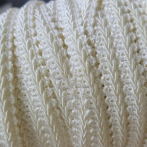 May include: A spool of off-white gimp trim featuring a decorative braided pattern with a raised central ridge and scalloped edges. The trim is tightly wound around a circular cardboard core, displaying a continuous length of textured fabric.