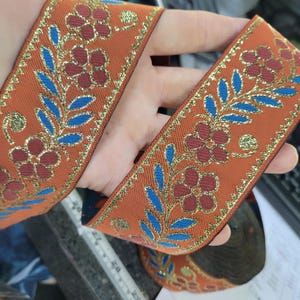 May include: Orange ribbon with a floral design. The ribbon features red flowers, blue leaves, and gold accents. The ribbon is likely used for crafts, sewing, or decorative purposes.