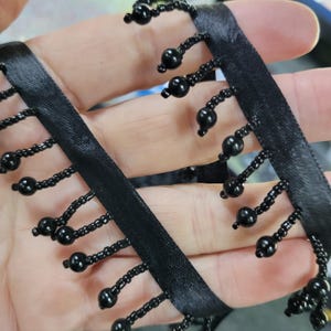May include: Black beaded fringe trim with a satin ribbon base. The trim features rows of small, shiny black beads hanging from the ribbon. This decorative trim is suitable for various craft and sewing projects.
