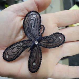 May include: A black butterfly-shaped applique with intricate lace detailing. The wings are outlined with black thread and adorned with small, sparkling rhinestones. The applique is designed with a delicate, airy aesthetic.