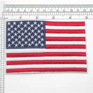 Könnte beinhalten: Ein gestickter amerikanischer Flaggen-Patch mit roten und weißen Streifen und einem blauen Sternenfeld. Der Patch ist etwa 6,5 cm breit und 9 cm hoch.