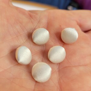 以下が含まれることがあります： オフホワイトの生地で覆われた、小さくて丸いボタンが5つ。 ボタンは滑らかで、サテンまたはシルクのような光沢があります。 手の中に配置され、サイズと質感が示されています。