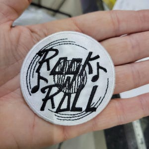 Könnte beinhalten: Ein runder, weißer, gestickter Aufnäher mit den schwarzen Worten "Rock and Roll". Das Design beinhaltet Noten und eine Schallplatten-Grafik in der Mitte. Der Aufnäher wird in einer Hand gehalten.