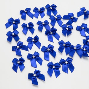 Pode incluir: Uma coleção de laços de cetim azul royal com centros de strass transparentes. Os laços estão dispostos em um padrão aleatório sobre um fundo branco.