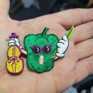 May include: Embroidered patch featuring a green bell pepper character wearing purple glasses and holding a violin and bow. The violin is yellow and purple. The pepper has a surprised expression.