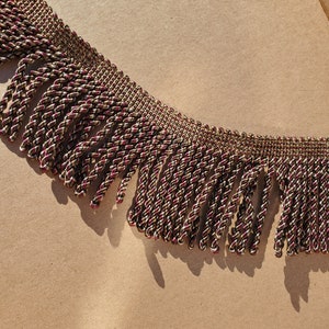 May include: A close-up of a brown and burgundy braided fringe trim. The fringe is made of a twisted cord and has a subtle sheen.