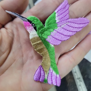 Puede incluir: Parche bordado de colibrí con cuerpo verde, alas moradas y un largo pico negro. El colibrí tiene garganta rosa y pecho dorado y blanco. Ideal para coser en ropa o proyectos de manualidades.