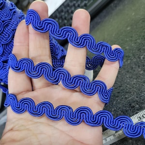 May include: A close-up of a vibrant blue decorative trim with a wavy, scroll-like design. The trim is held in a hand, showcasing its texture and detail. This trim is ideal for sewing, crafting, and adding a decorative touch to various projects.