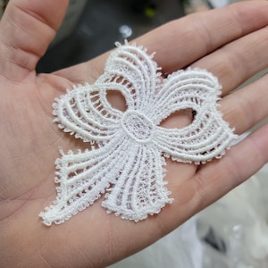 May include: A white lace bow applique with intricate detailing. The bow features a central oval shape and symmetrical, curved lines, creating a delicate and elegant design. The edges are scalloped, adding a refined touch to this decorative element.