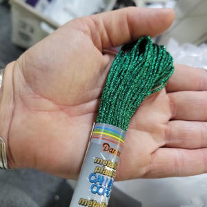 May include: A bundle of green metallic plastic canvas cord held in a hand. The cord is tightly wound and has a shimmering, glittery appearance. The label reads "Dance" and "metallic plastic canvas cord".