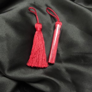 Puede incluir: Dos adornos de borla roja sobre un fondo de satén negro. Las borlas están hechas de hilos rojos finos y tienen un lazo en la parte superior para colgar.
