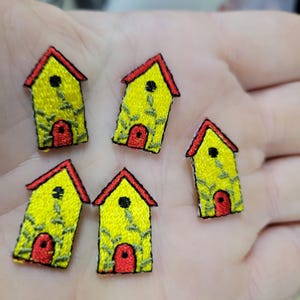 Könnte beinhalten: Fünf gestickte Hausaufnäher. Jeder Aufnäher zeigt ein gelbes Haus mit rotem Dach und roter Tür. Grüne Stickdetails verzieren das Haus. Die Aufnäher sind klein und detailliert, ideal zum Basteln oder Verzieren.