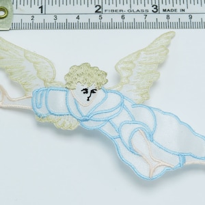May include: A white and blue embroidered angel with wings, a halo, and a flowing robe. The angel is flying with its arms outstretched. The angel is approximately 2 inches tall.