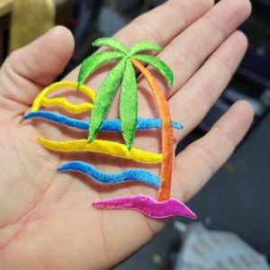 May include: Embroidered patch featuring a tropical beach scene. The design includes a green palm tree, a yellow sun, and blue and yellow waves. The patch is made with colorful thread and has a detailed, textured appearance.