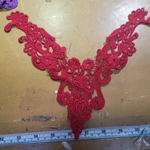 May include: A vibrant red lace applique with an intricate floral and leaf design. The applique is symmetrical, with a central pendant shape and extending, curved arms. The delicate lace is likely for embellishing clothing or craft projects.