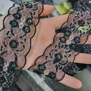May include: Black lace trim with a scalloped edge, featuring silver embroidery and iridescent sequins. The trim has a floral design and is held up by a hand. This trim is suitable for sewing and crafting projects.
