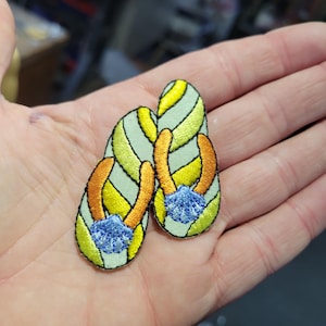 May include: Embroidered flip-flop patches featuring a light green base with yellow and orange accents. Each patch has a blue seashell detail. The patches are designed with a stained-glass effect.