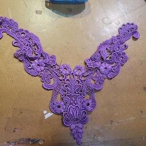 May include: A purple lace applique with a floral and scroll design. The applique is in a V-shape, with detailed openwork and a scalloped edge. It is likely used for embellishing clothing or crafts.