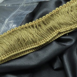 May include: A close-up of a gold-colored fringe trim with a woven edge. The trim is lying on a black fabric surface.