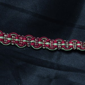 May include: A close-up of a red and green braided trim with a repeating pattern of rounded shapes. The trim is on a dark blue fabric background.