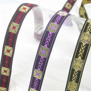 Op de afbeelding: Close-up van decoratief lint in zwart met geometrische patronen in goud, rood en paars. Het lint wordt waarschijnlijk gebruikt voor knutselen, naaien of het verfraaien van diverse items. Het ontwerp bevat vierkanten en pijlvormige figuren.