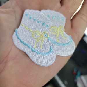May include: White embroidered baby booties with blue and yellow accents. The booties feature a scalloped edge and bows. The embroidery is detailed, creating a delicate and charming design. Ideal for baby showers or crafting projects.