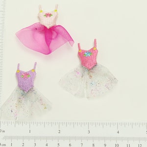 May include: Three miniature ballerina dresses with tulle skirts. The dresses are pink, purple, and white with embroidered flowers. The skirts are layered and have a sparkly finish.