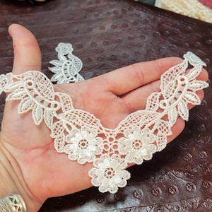 May include: Ivory-colored lace applique with a floral design. The applique features a V-neck shape with three prominent flower motifs and delicate leaf patterns. A smaller, matching lace piece is also visible.