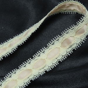 May include: A close-up of a pale yellow lace trim with a row of oval-shaped pink satin accents. The trim is on a black fabric background.