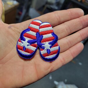 May include: Embroidered patch of two flip-flops with a patriotic design. The flip-flops feature red and white stripes, blue straps, and a white star. The patch has a blue border.