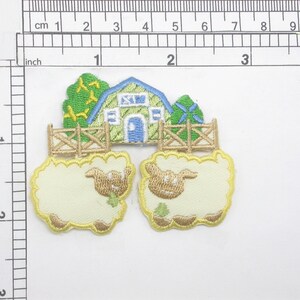 May include: Two yellow embroidered sheep with brown faces and green leaves in their mouths, standing in front of a green and blue embroidered farmhouse with a brown fence.