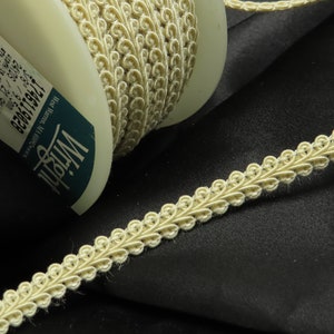 May include: A spool of off-white trim with a decorative, scalloped edge. The trim is wound around a white spool with a blue label that reads "Wright" and "West Warren, MA 1746119028".