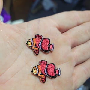 May include: Two embroidered clownfish patches. Each patch features a vibrant orange and red color scheme with black outlines. The fish have detailed fins and a distinctive white stripe, perfect for adding a nautical touch to clothing or accessories.