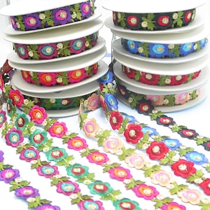 Peut inclure: Plusieurs bobines de ruban floral décoratif de différentes couleurs, dont le violet, le rose, l'orange, le vert, le bleu et le rouge. Chaque ruban présente des fleurs brodées avec des accents dorés et des feuilles vertes.